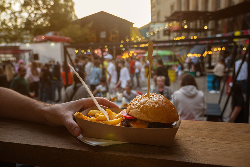 A burger and fries sitting in a cardboard tray. A hand holds onto the tray. Both rest on a wooden table. In the blurred background is a crowd of people standing around outside.
