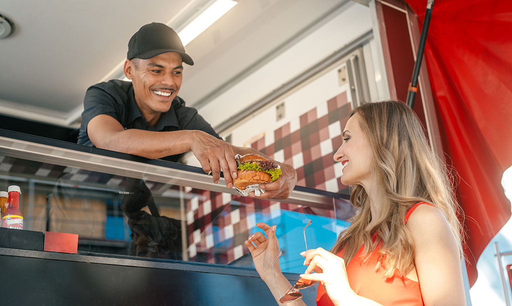 A smiling man in a food truck handing a burger to a smiling woman who stands outside of the food truck.